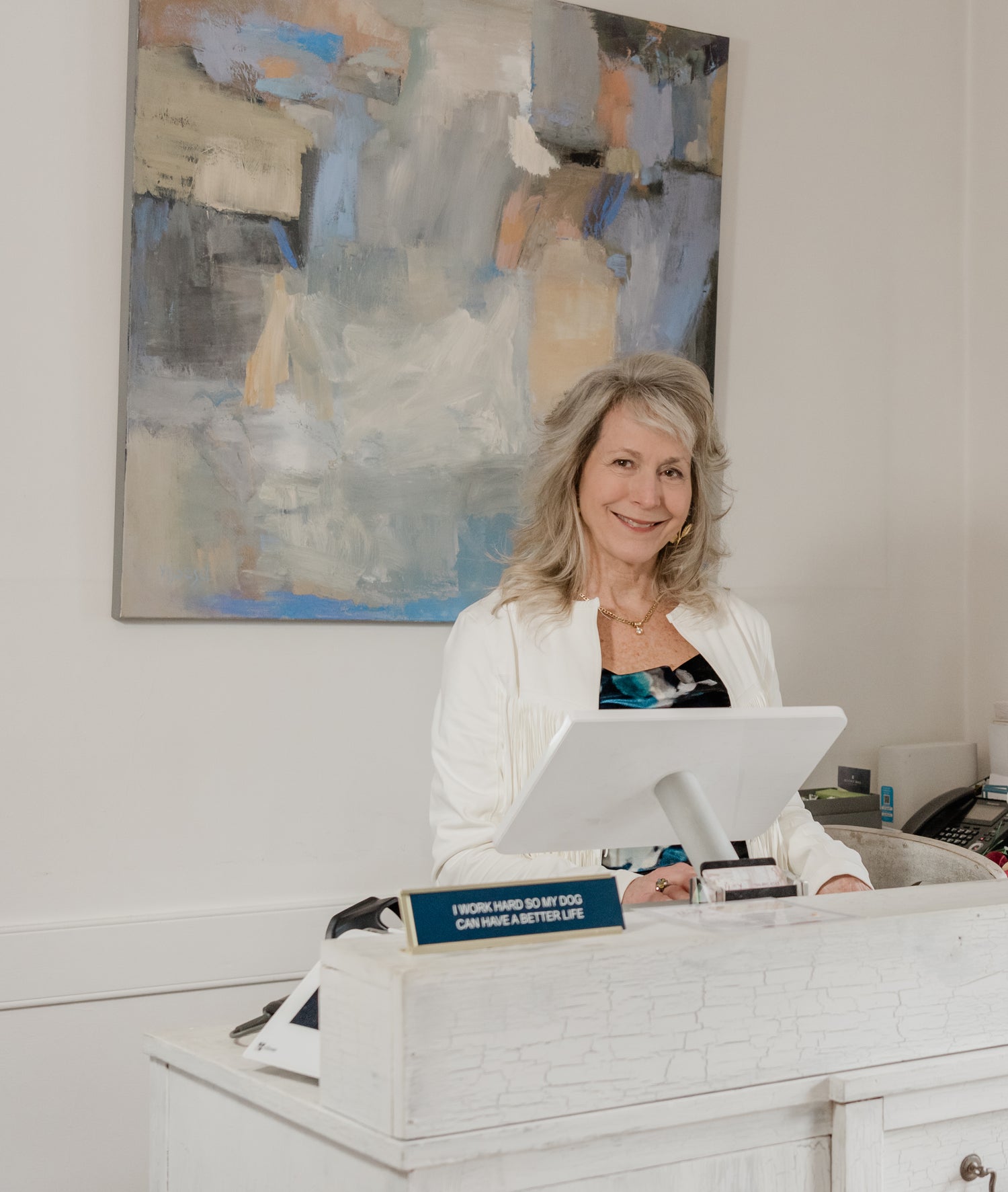 Lori Savio sitting at the check out area of Home, Heart & Soul next to a blue desk sign that says, "I work hard so my dog can have a better life."