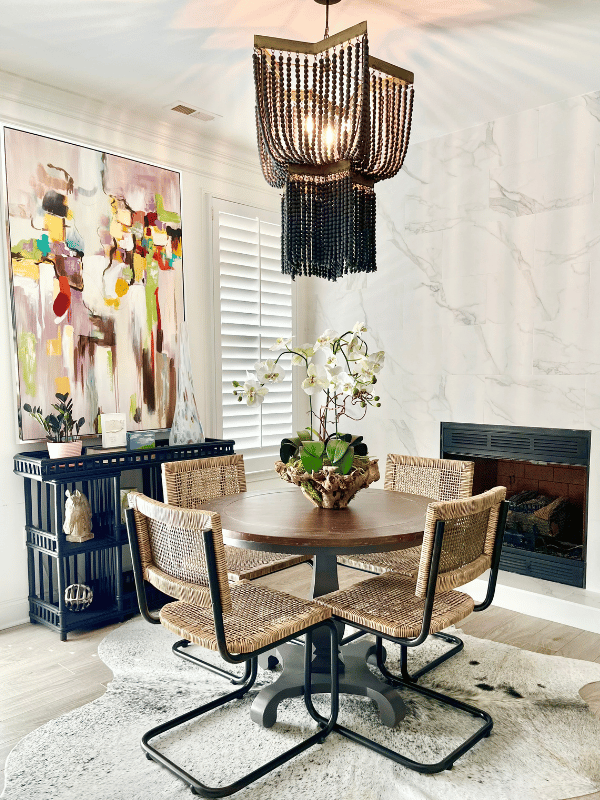 Photo of a breakfast nook with beautiful marbled wall and fireplace, bright abstract art, a circular wooden table with four chairs with wicker accents, and stunning chandelier lighting in the shape of a star with intricate beadwork.
