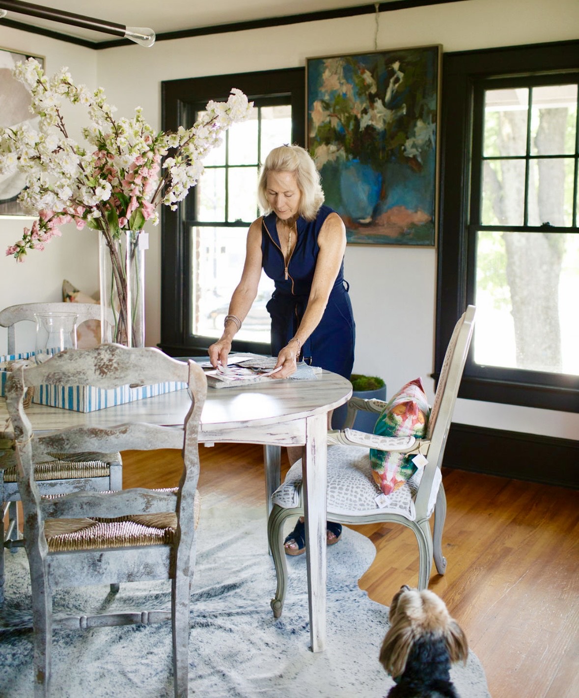 Lori Savio, interior designer, in a blue dress standing at a dining table with her dog nearby, in a room with large windows and a painting on the wall.