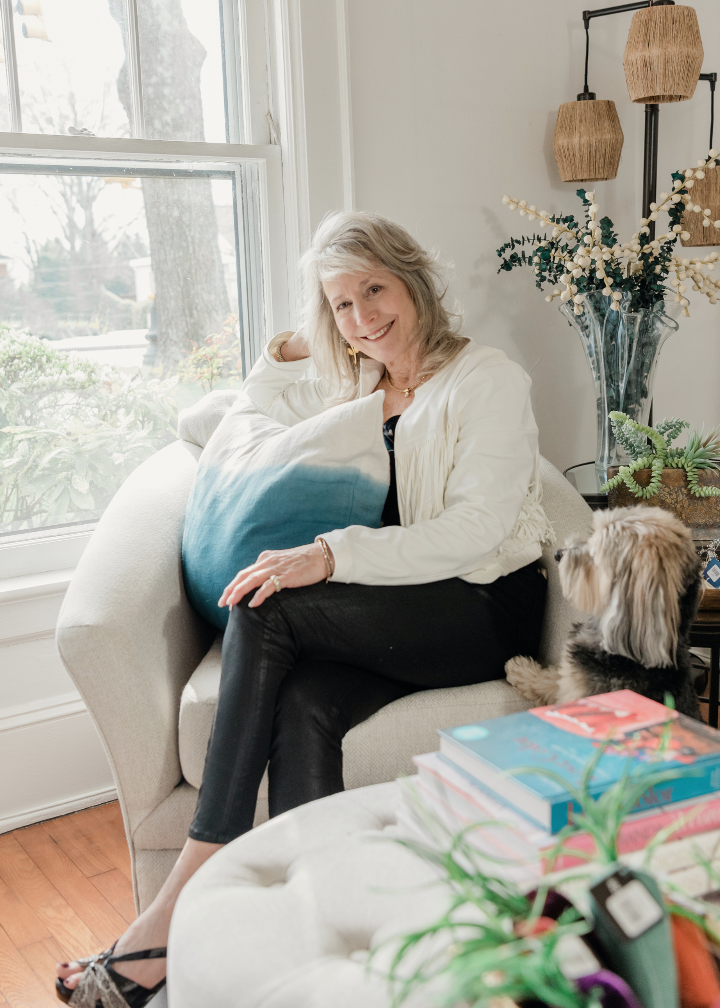 Lori Savio, Interior Designer and Owner of Home, Heart & Soul, sitting on a beige chair by a window in her home décor shop