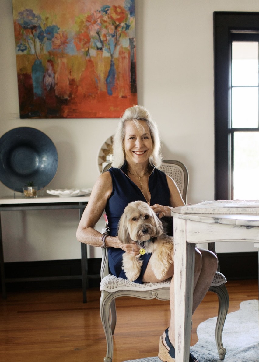 Lori Savio sitting at a table with her dog, smiling, in a room with a colorful painting on the wall.